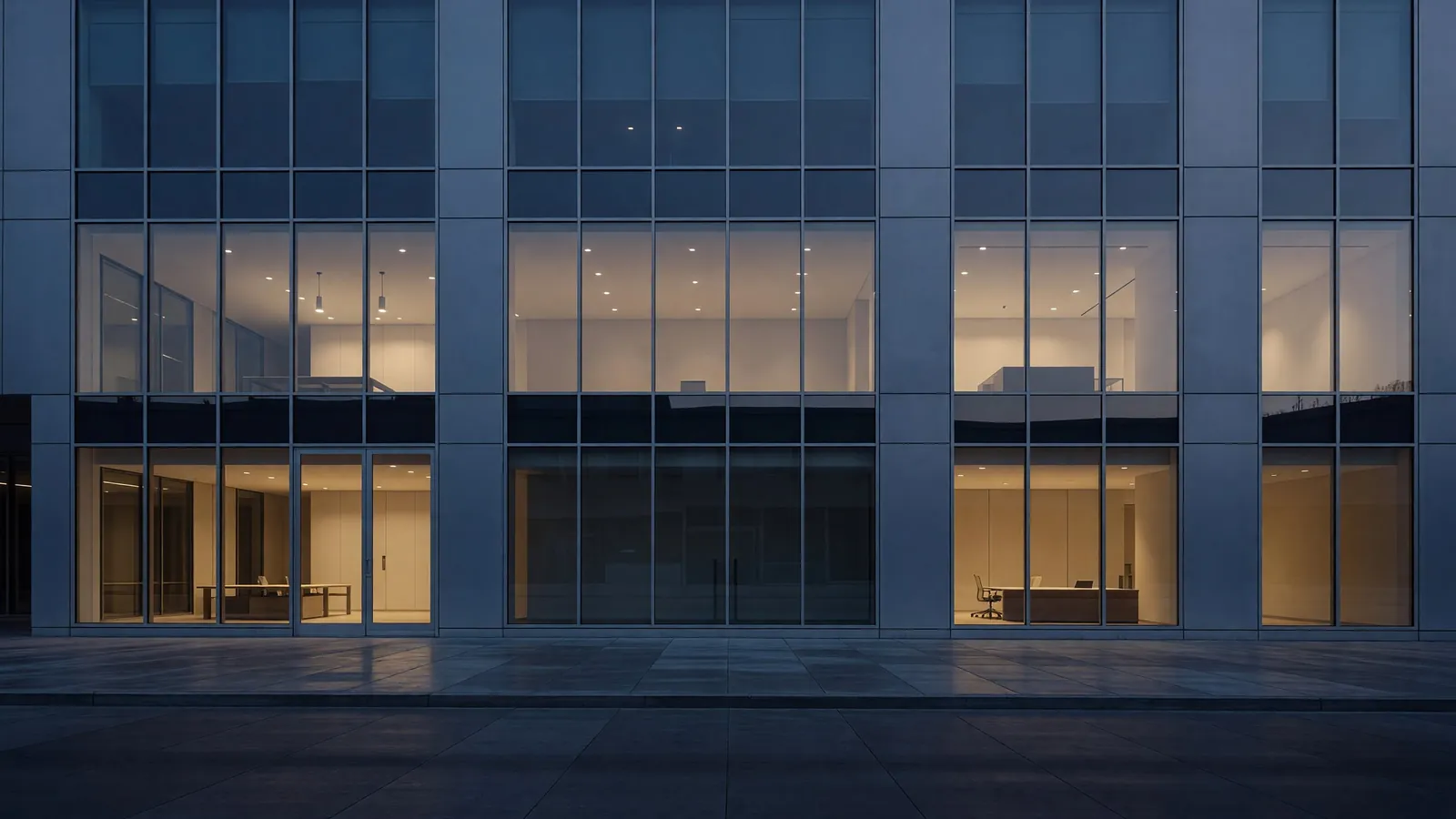 Modern commercial office building at dusk with warm light glowing through floor to ceiling windows onto an empty sidewalk.