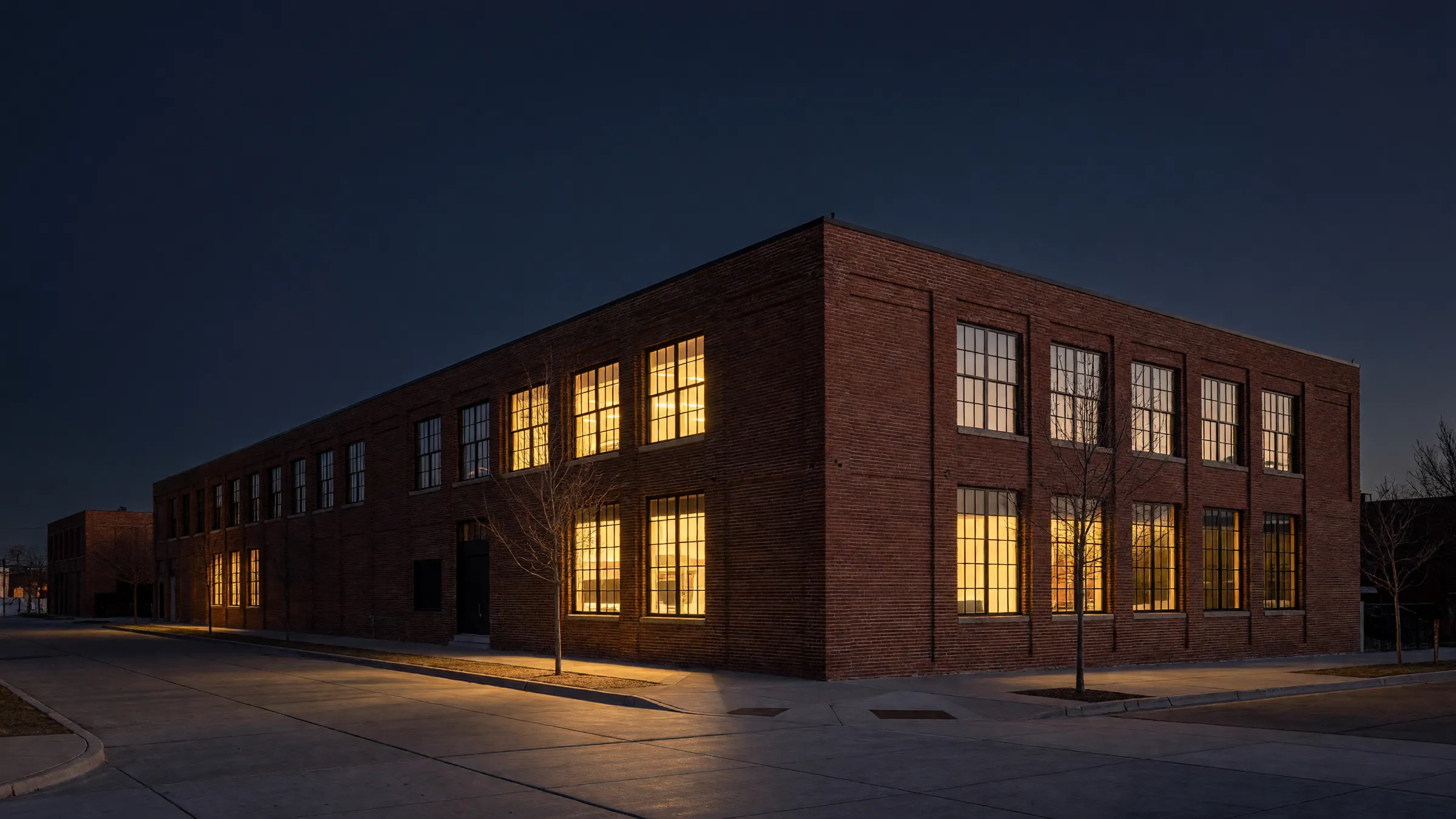 A converted red brick warehouse office building in Denver at dusk, its tall windows lit warm above an empty sidewalk